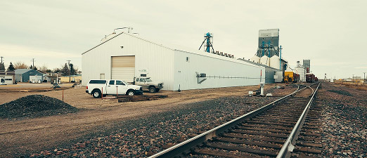 A white industrial building stands next to a set of railroad tracks, with vehicles parked outside and silos or grain elevators visible in the background under an overcast sky.