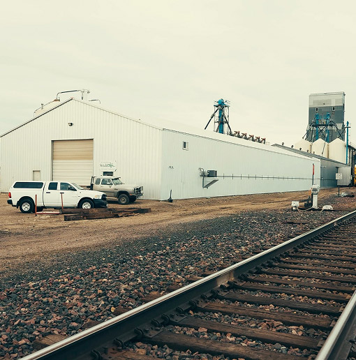 A white industrial building stands beside railroad tracks. Two white trucks are parked outside the building on a gravel lot under an overcast sky. Industrial equipment is visible in the background.
