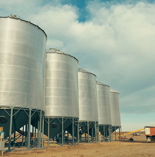 Four large, silver metal grain silos stand in a row on a farm under a partly cloudy sky, with open land and farming equipment visible in the background.