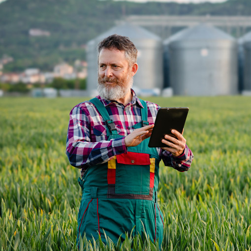 A bearded man in plaid shirt and overalls stands in a green field, using a tablet. Large silos and a hilly landscape are visible in the background.