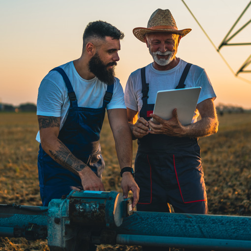 Two farmers in overalls stand in a field at sunset, one holding a tablet and the other adjusting machinery. They are smiling and appear to be discussing or reviewing information together.